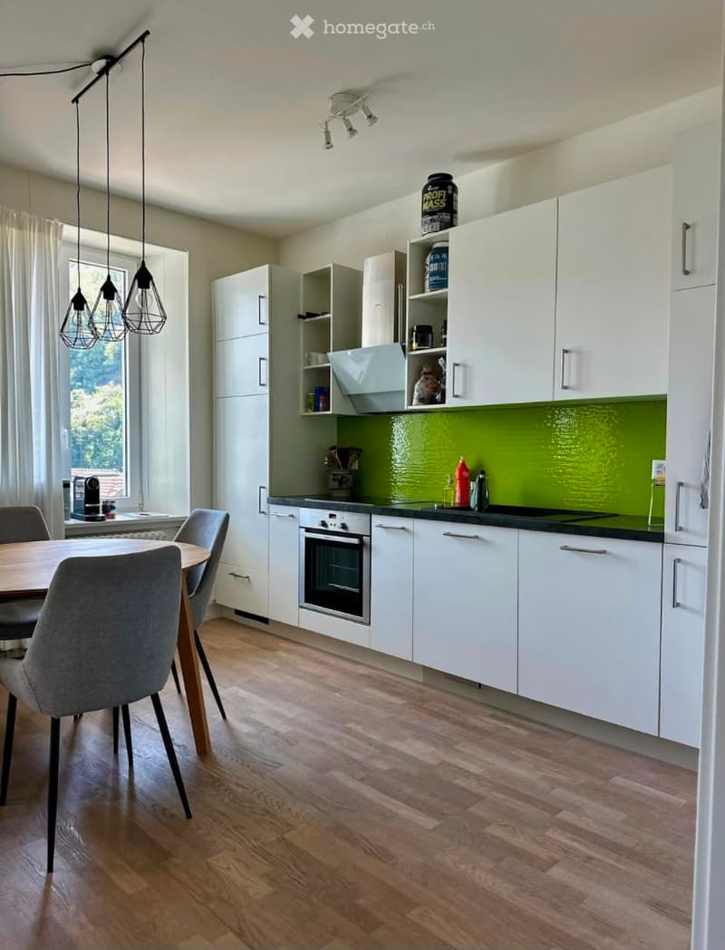 Modern kitchen with white cabinets, a green backsplash, and a wooden dining table with chairs. Features an oven, countertop, and pendant lighting.
