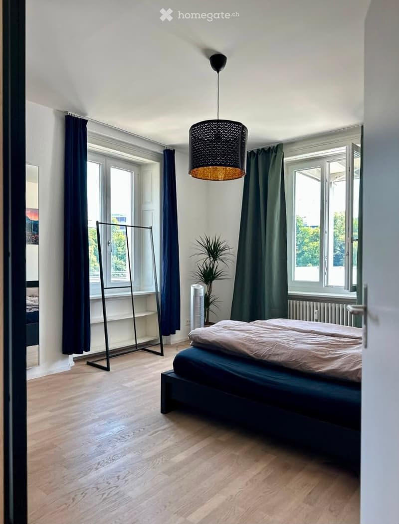 Modern bedroom with wooden flooring, large windows, dark curtains, a bed with a beige duvet, a hanging lamp, a plant, and a wardrobe.