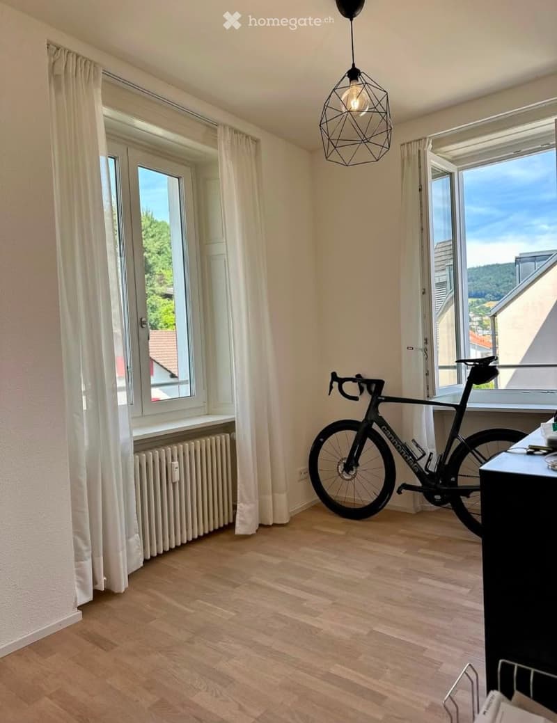 Empty room with wooden flooring, large windows with white curtains, radiator, and a bicycle placed near the window.