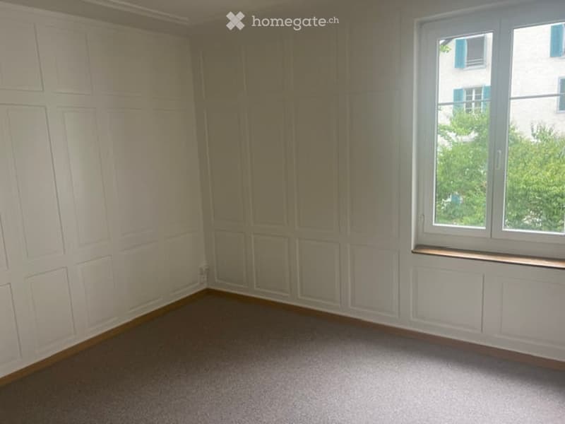 Empty room with white paneled walls, carpeted floor, and a large window with a view of greenery.