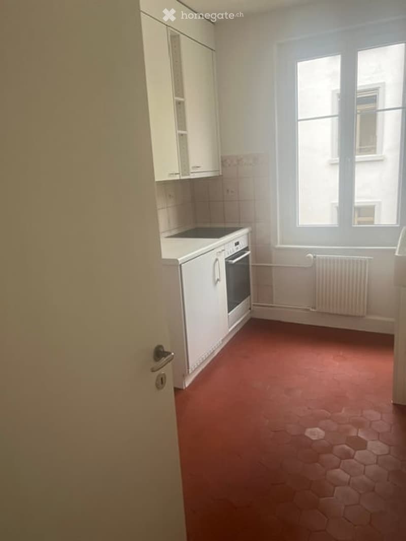 Empty kitchen with white cabinets, a stove, and a radiator under the window. The floor is red hexagonal tiles.