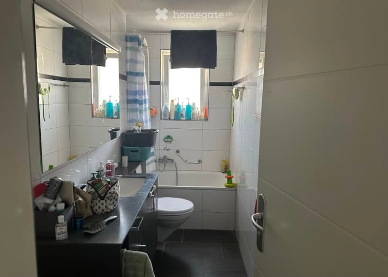 Modern bathroom with white tiles, a large mirror, a sink with various toiletries, a toilet, a bathtub, and a shower area. The space is well-lit with natural light coming through a window, and towels are neatly hung on the walls.