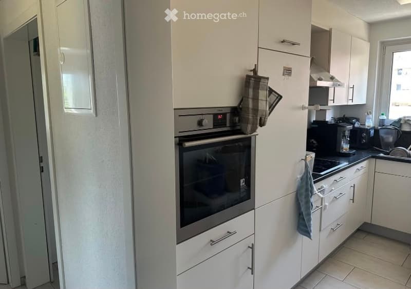 Modern kitchen with white cabinets, stainless steel oven, coffee maker, toaster, and various kitchen appliances. A dish rack is visible, and a plaid oven mitt hangs on the cabinet.