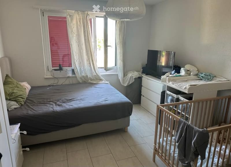 A well-lit bedroom with a bed, pillows, a wooden cradle, a wardrobe, and a television. The room features tiled flooring and a window with white curtains.