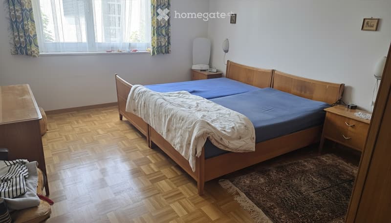 A furnished bedroom with a wooden bed, blue bedding, a nightstand with a lamp, a small table, and a chair near the window.