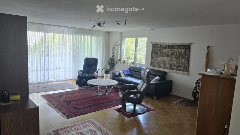 Living room with hardwood flooring, large windows with sheer curtains, a black leather sofa, a round coffee table, a black leather chair, and a floor lamp. The room is decorated with several rugs and a large tapestry on the wall.