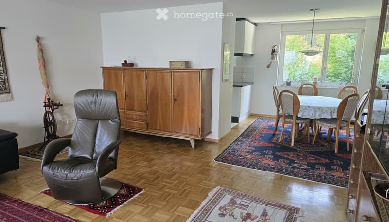 A spacious, well-lit living and dining area with wooden flooring, a large leather sofa, a wooden cabinet, a dining table with chairs, and a small kitchen area with cabinets and appliances.