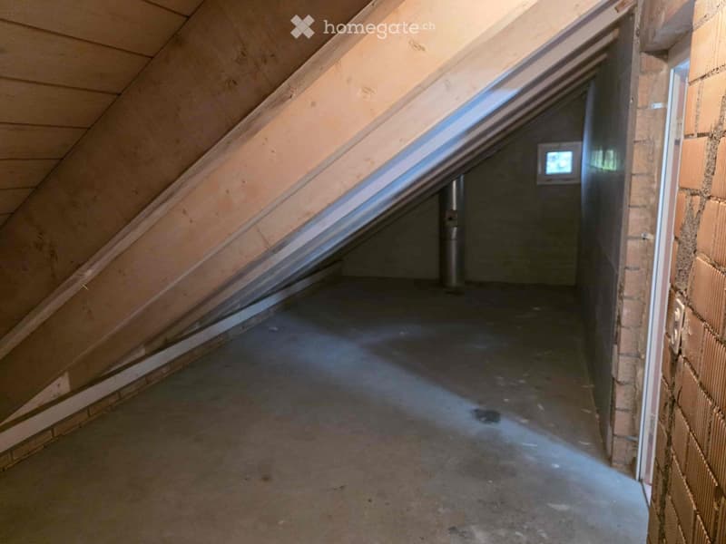 The image shows an attic space with exposed wooden beams and a slanted ceiling. The floor appears to be concrete or a similar material, and there is a small window visible in the wall. The space seems to be unfinished and used for storage or utility purpo