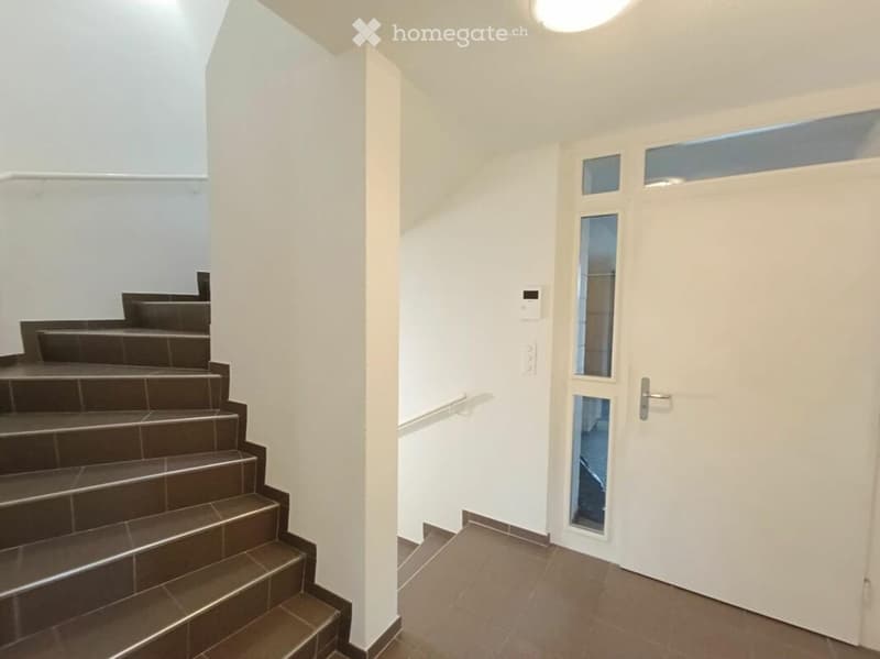 Empty hallway with white walls, tiled floor, staircase leading upwards, and a white door with glass panels on the right side.