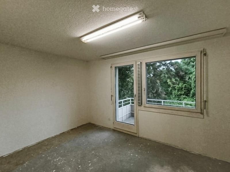 Empty room with beige walls, concrete floor, large window, and sliding glass door leading to a balcony.