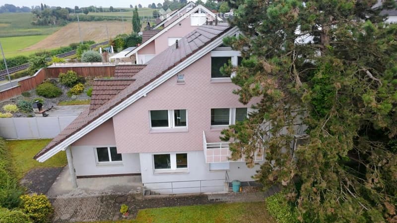 A two-story house with a pink exterior and white trim, featuring multiple windows, a balcony, and a well-maintained garden.