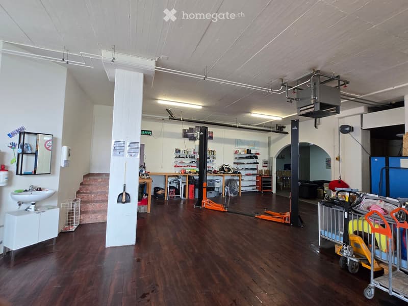 A spacious, well-lit room with wooden flooring, a sink, shelves, a table, and various equipment including a cart and a stool.