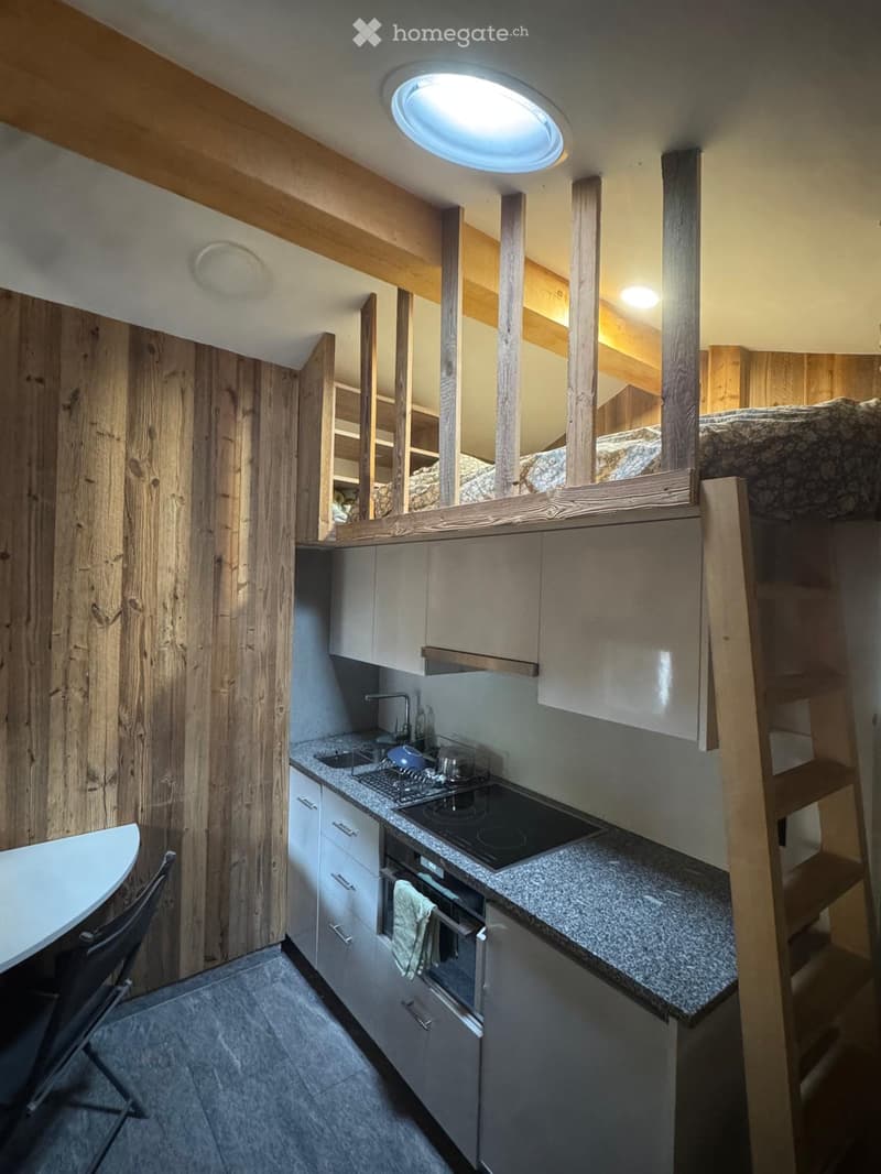 Modern kitchen with granite countertops, stainless steel appliances, and wooden beams. Features a sink, stove, oven, and a small dining area with a chair and table.