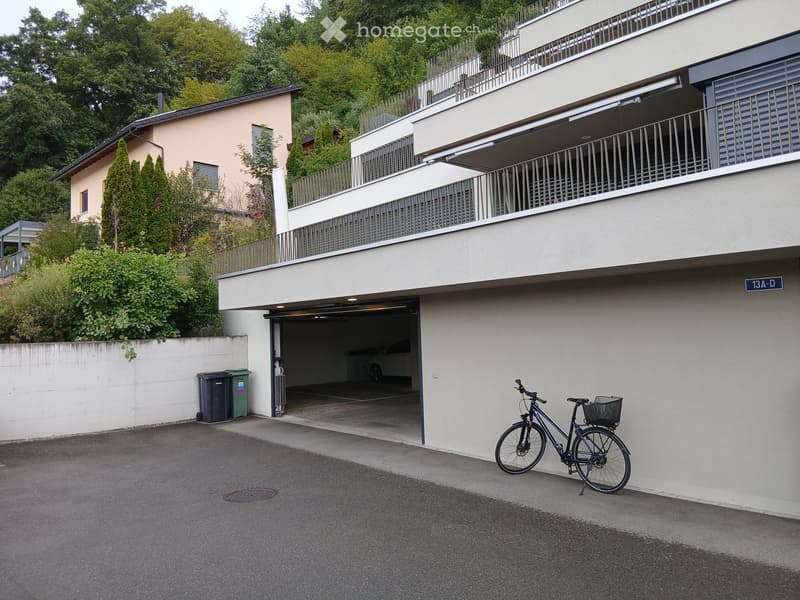 Modern multi-story building with a garage entrance, bicycle parked outside, and greenery in the background.