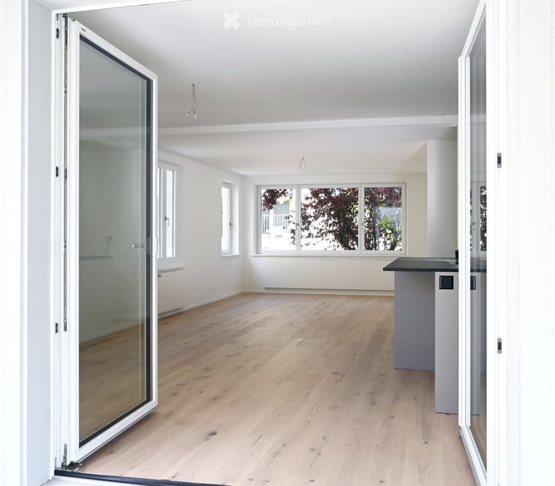 Empty room with light wooden flooring, white walls, large windows, and a sliding glass door leading outside.