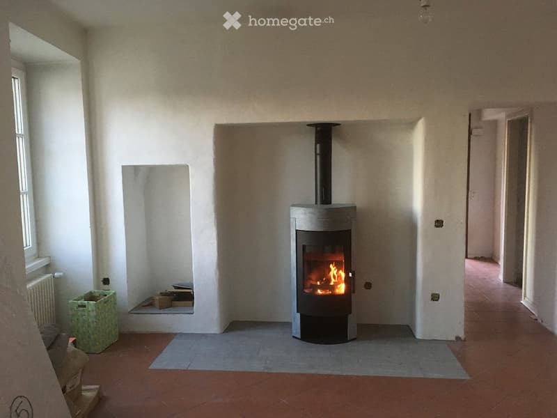 Empty room with a modern wood-burning stove, tiled floor, and white walls. The room has a window on the left and a doorway on the right.