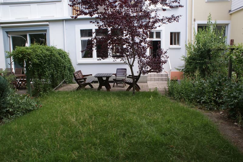 Garden with a well-maintained lawn, small patio area with wooden chairs and a table, surrounded by shrubs and trees, with a house in the background featuring white walls and multiple windows.