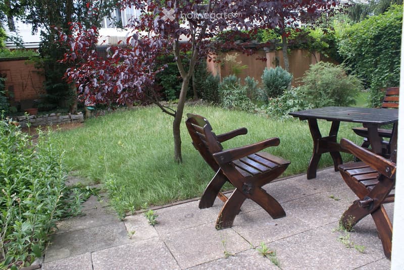 A well-maintained garden with a wooden table and chairs on a paved patio, surrounded by green grass, trees, and shrubs.