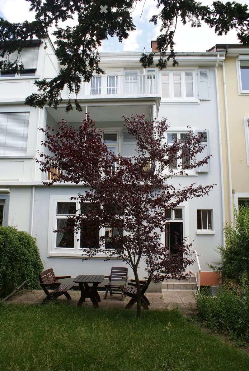 Garden area with a small tree, wooden table, and chairs in front of a two-story house with white walls and multiple windows.