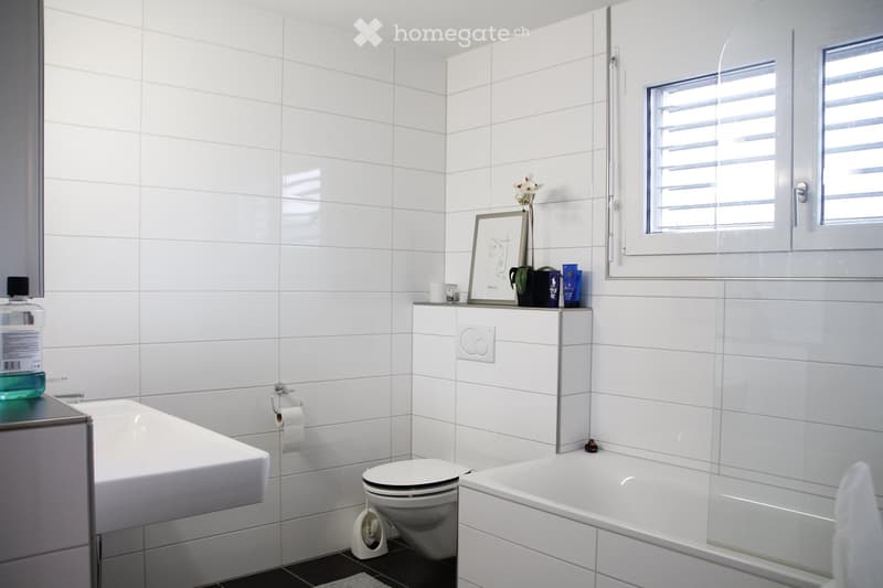 Modern bathroom with white tiled walls, a bathtub, toilet, sink, and a framed picture on a shelf above the sink.