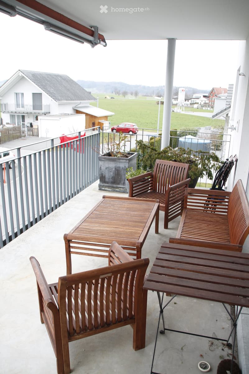 Balcony with wooden furniture including a table and chairs, overlooking a rural landscape with houses and fields.