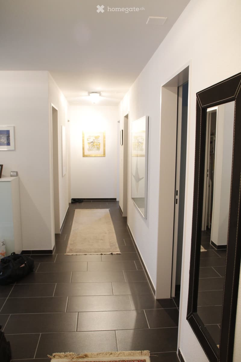 A modern hallway with dark tiled flooring, white walls, and several doors leading to different rooms. There is a small rug in the center and a mirror on the right wall.