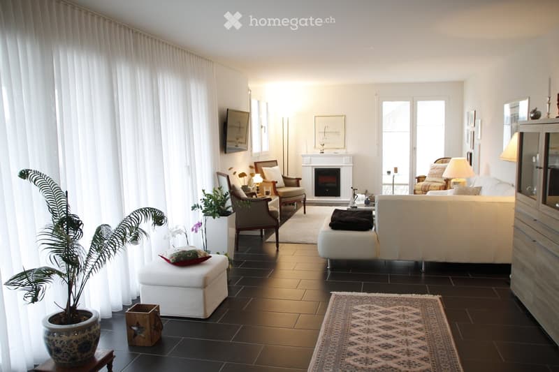 Modern living room with dark tiled flooring, white walls, large windows with white curtains, fireplace, sofa, ottoman, armchairs, lamps, and decorative plants.