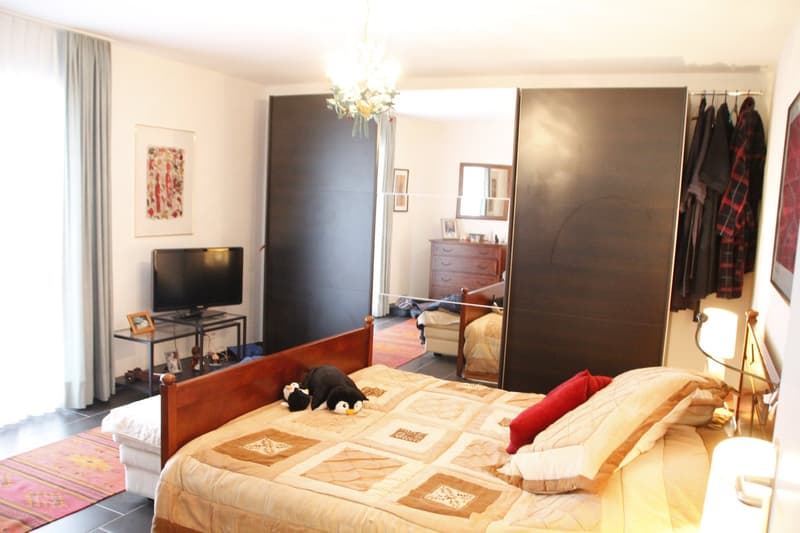 A well-furnished bedroom with a large bed, nightstand, television, mirror, wardrobe, dresser, chandelier, and picture frames. The room features a cozy quilted bedspread and decorative pillows.