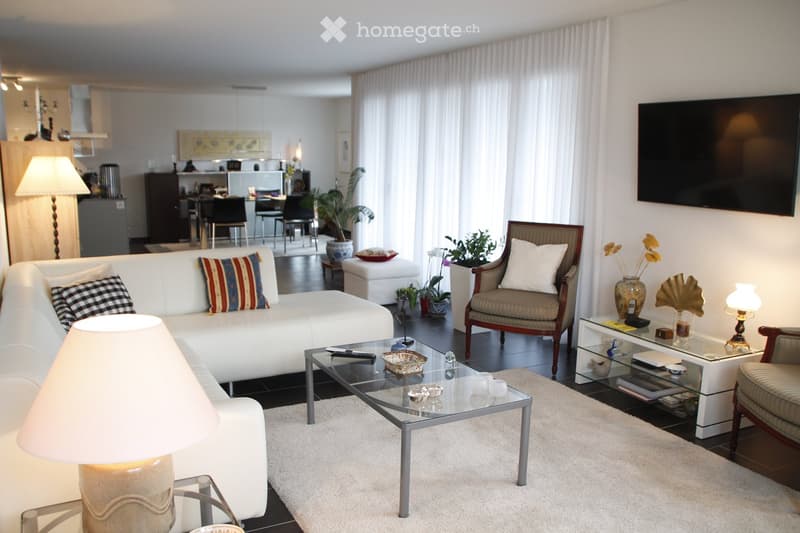 Modern living room with white sofa, glass coffee table, armchair, side table, and decorative items. The space features large windows with white curtains and a view of a dining area in the background.