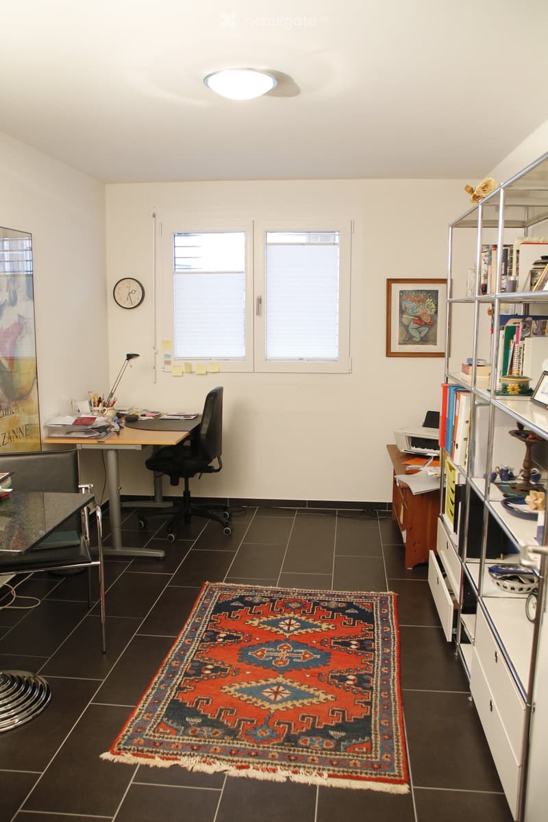 Office space with a desk, chair, shelf, lamp, clock, and a colorful rug on the floor.