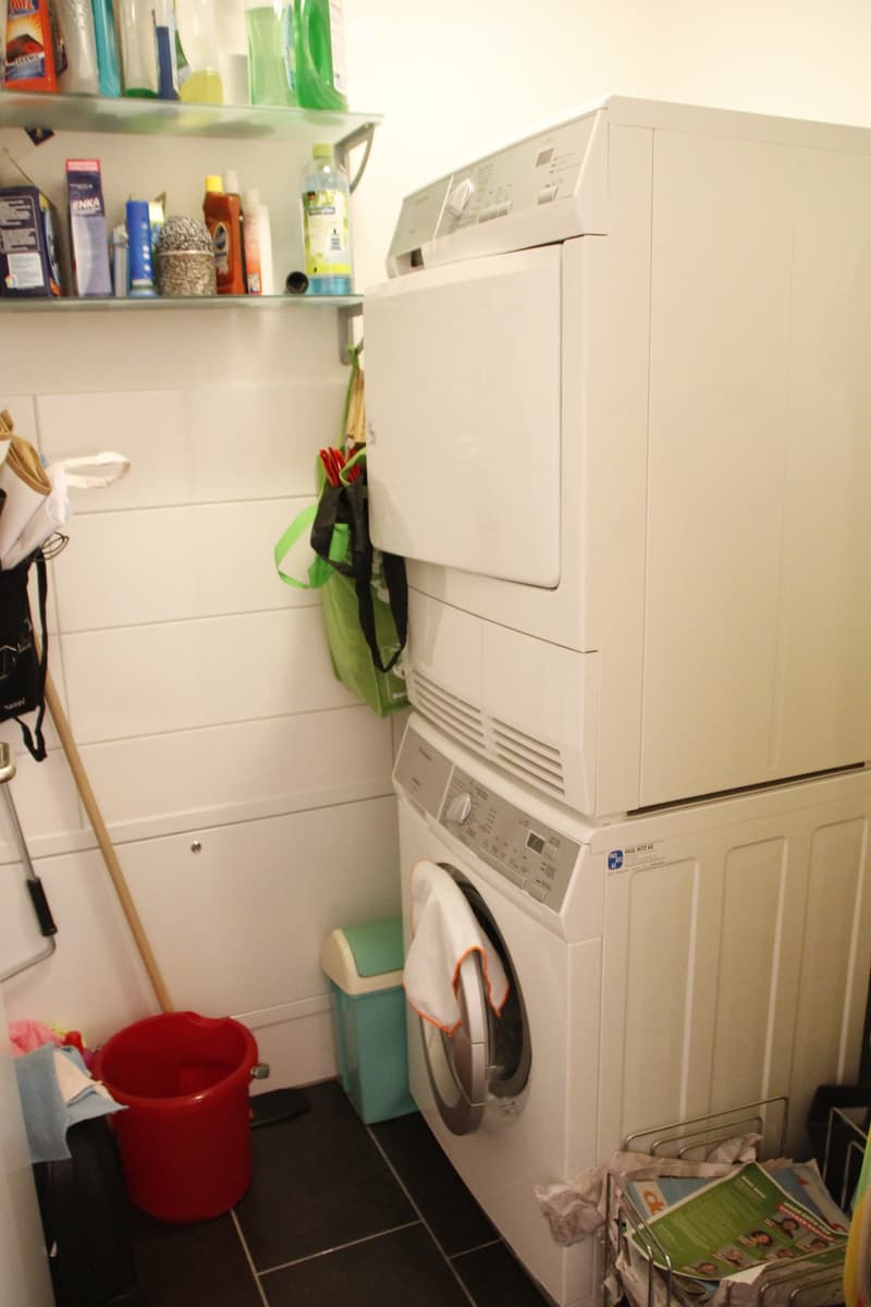 A small bathroom with a white washing machine, shelves with cleaning supplies, a red bucket, and a towel hanging on the machine.