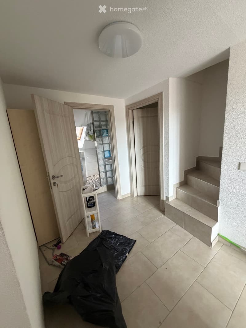 Empty hallway with tiled floor, white walls, and a staircase leading upwards. A black sheet is on the floor, and a small shelf with cleaning supplies is visible near an open door.