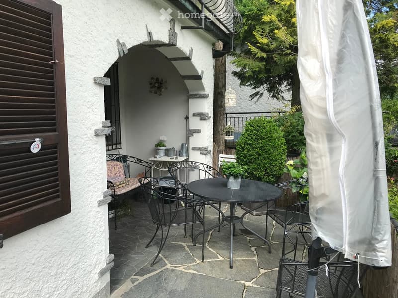 Outdoor patio with stone flooring, a round black metal table, several black metal chairs, a small bench, and potted plants. A large white umbrella is partially visible on the right side.