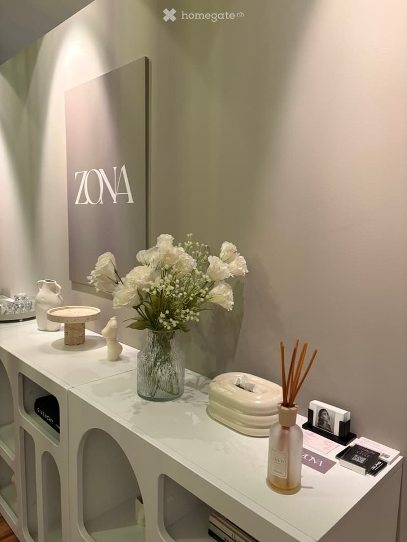 A modern, minimalist vanity area with a white table, a vase of white flowers, a decorative bottle, a mirror, and various small decorative items.