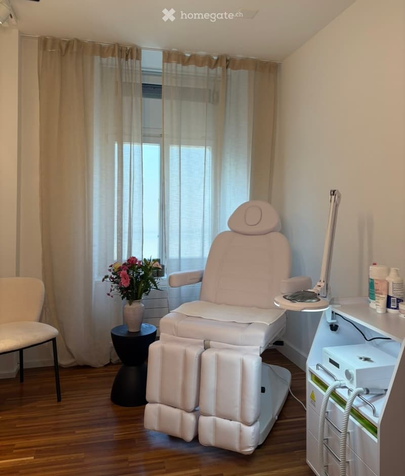 A well-lit room with a large, plush white massage or treatment chair, a small round black table with a vase of pink flowers, a white cabinet with medical or cosmetic equipment, and a small white chair near the window.