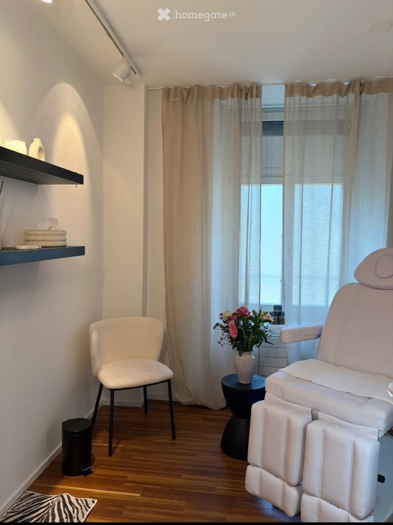 A well-lit room with wooden flooring, a large window with curtains, a white massage chair, a small round table with a vase of flowers, a white chair, and a shelf with decorative items.