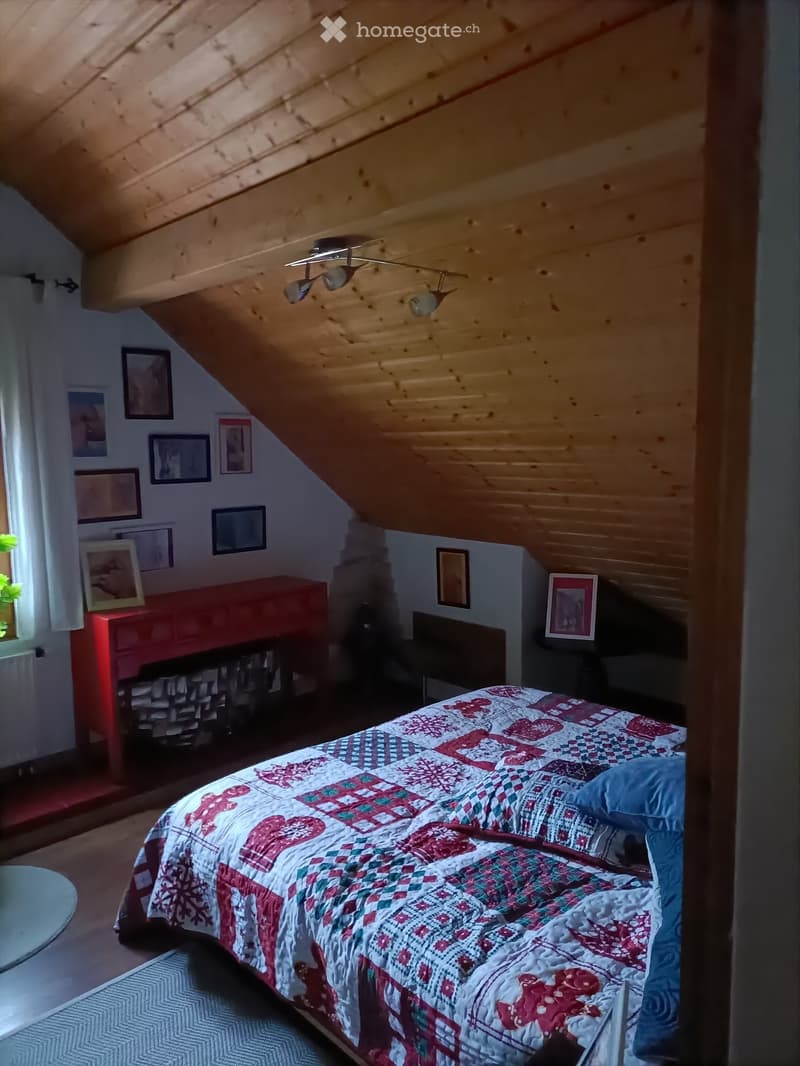 Cozy attic bedroom with wooden slanted ceilings, a bed with a colorful quilt, a nightstand, a mirror, and framed pictures on the walls.