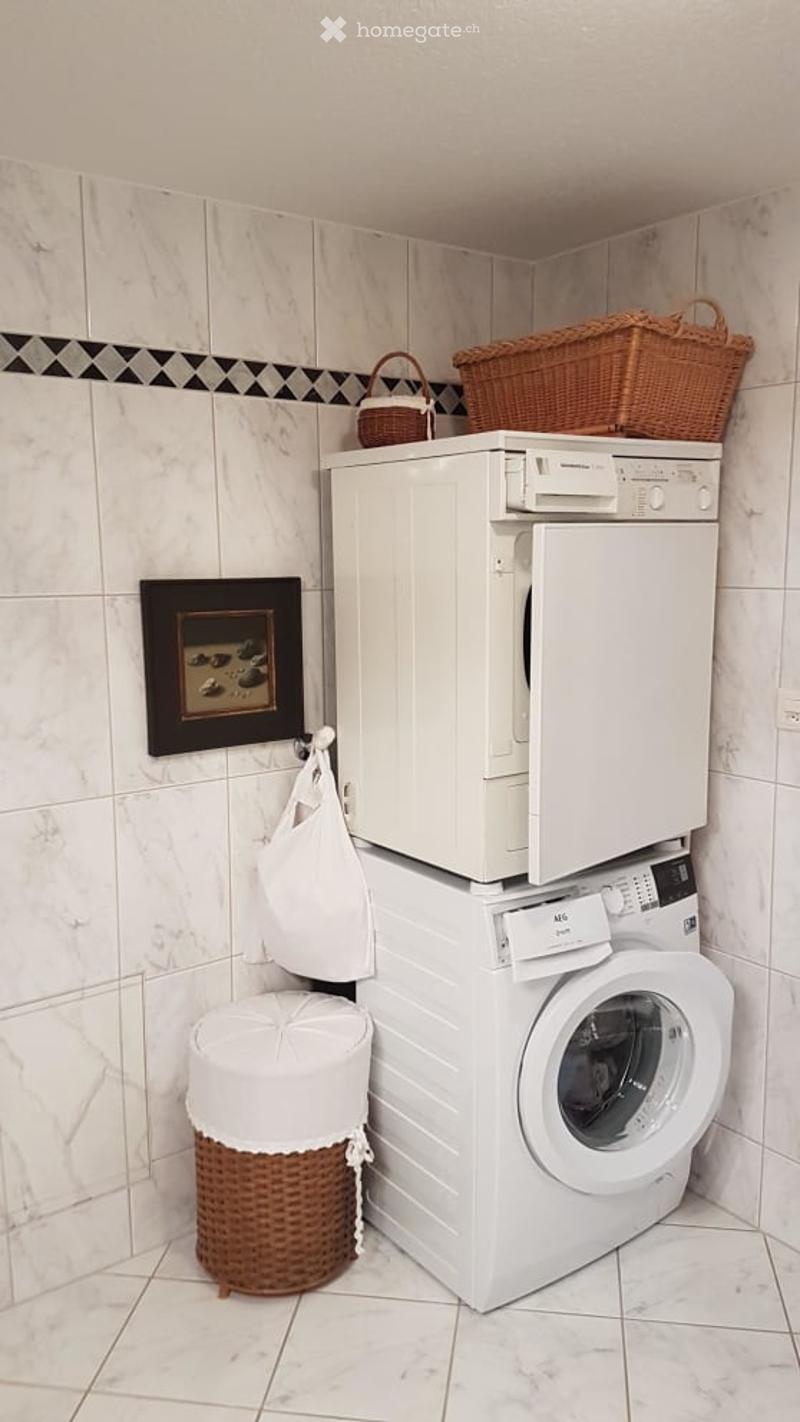 A compact bathroom with a stacked washing machine and dishwasher unit, wicker baskets on top, a small stool, and a framed picture on the wall.