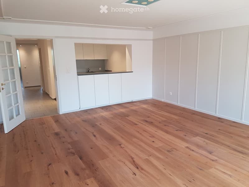 Empty room with light wooden flooring, white walls, and a small kitchen area visible through an open doorway.