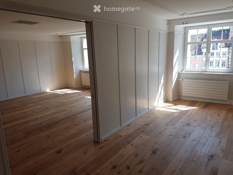 Empty room with light wooden flooring, white walls, and large windows allowing natural light. Radiators are visible along the walls.