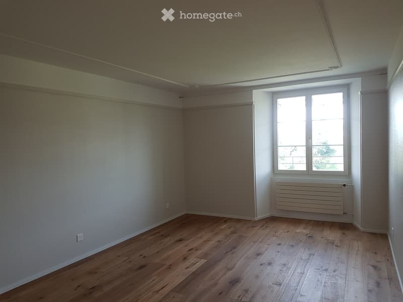 Empty room with wooden flooring, white walls, and a large window with a radiator below it.