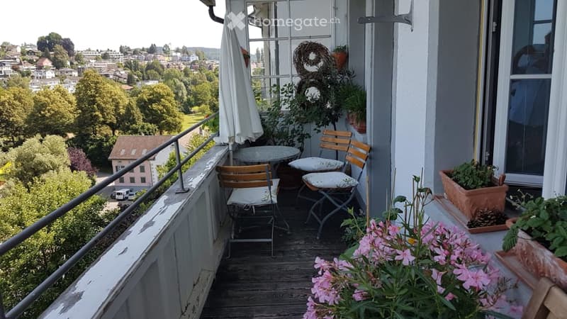 Balcony with wooden floor, metal railing, small round table, two wooden chairs, potted plants, and a view of the city and greenery.