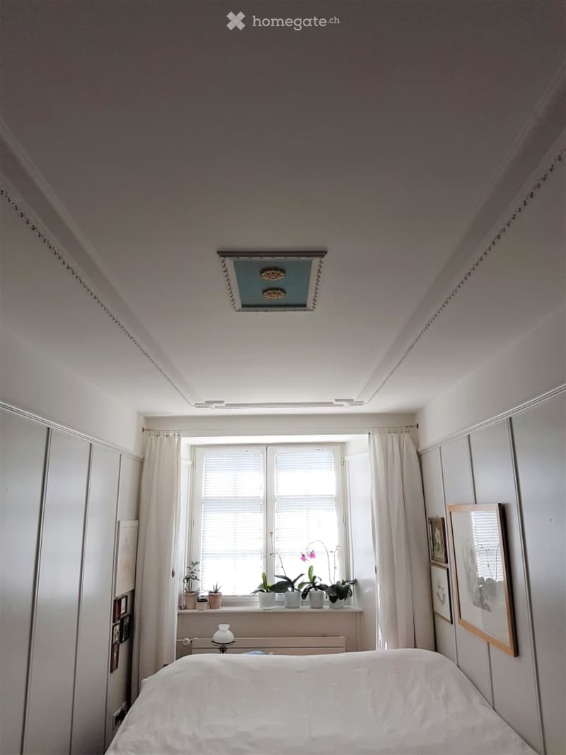 Bedroom with a bed, a window with curtains, a small table lamp, and several potted plants on the windowsill. The room has white walls and a decorative ceiling.