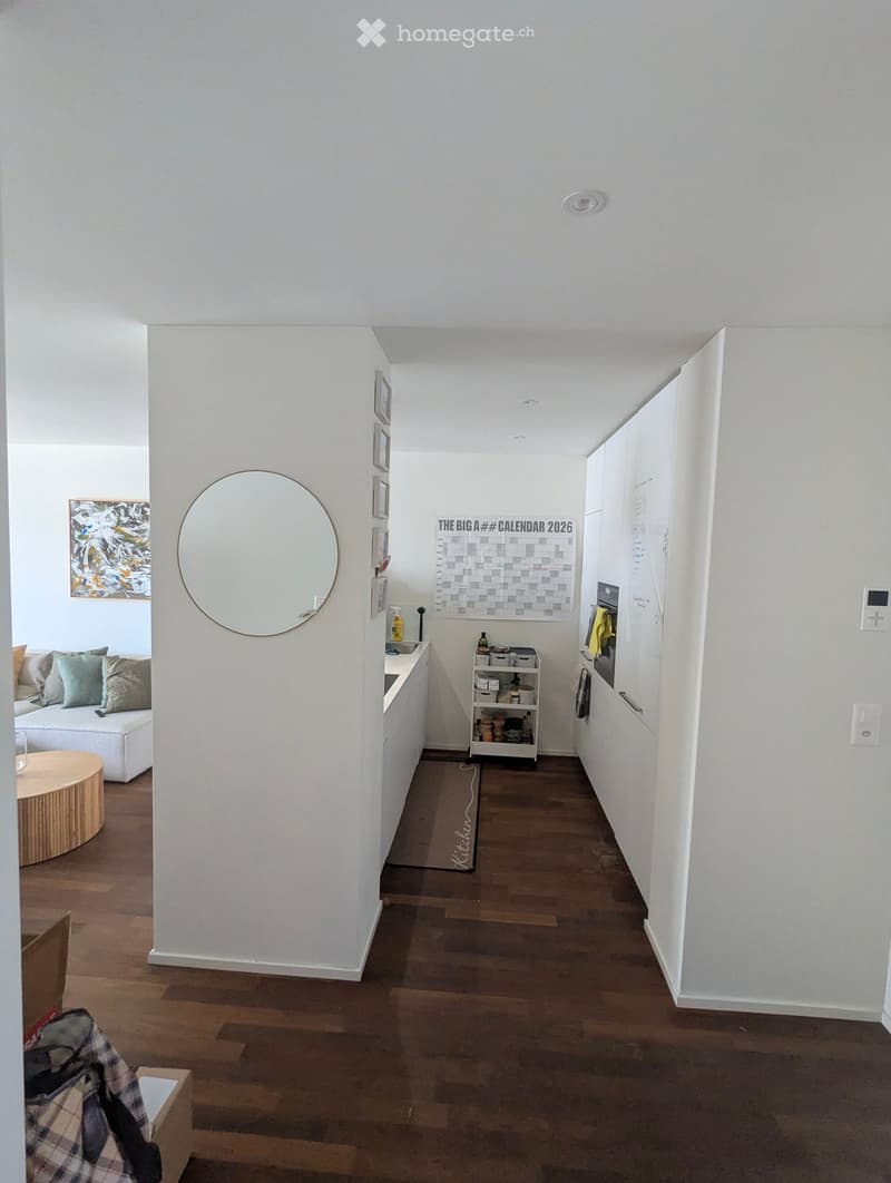 A modern hallway with white walls, wooden flooring, and a round mirror on one wall. A small kitchen area is visible to the right, with cabinets, a countertop, and a small shelf with kitchen items. A calendar and framed artwork are on the wall, and a small