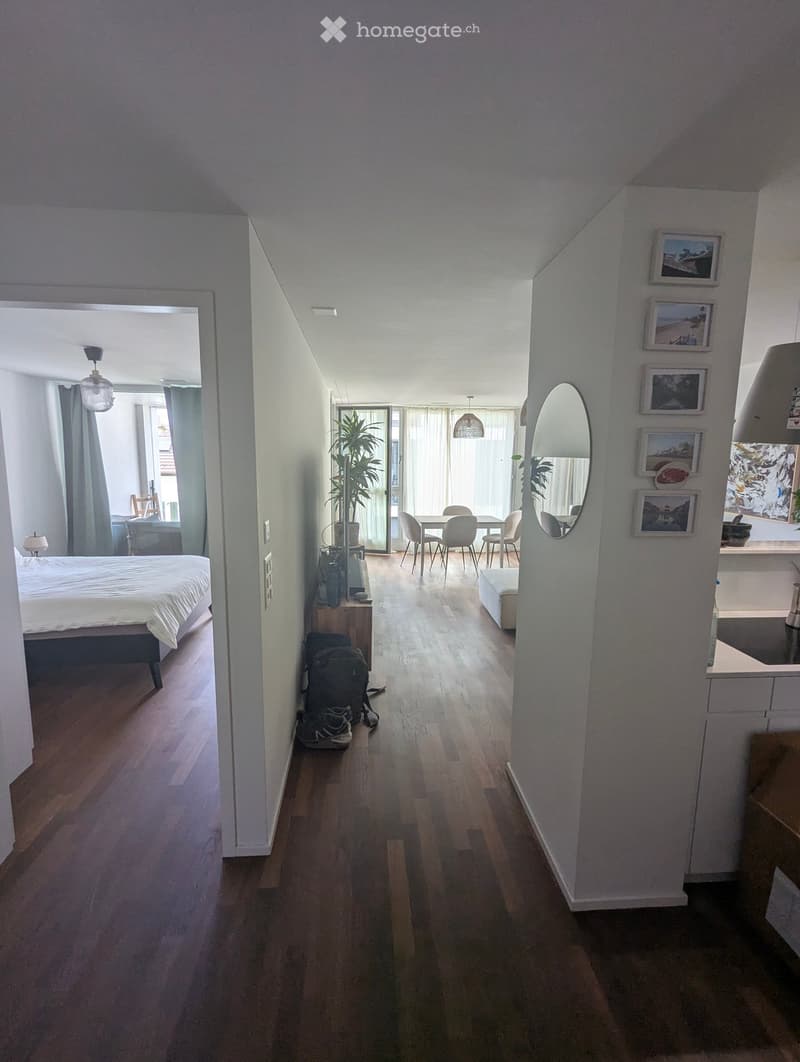 A modern hallway with wooden flooring, leading to a bedroom and a dining area. Features include a mirror, framed pictures, and a small plant in the corner.
