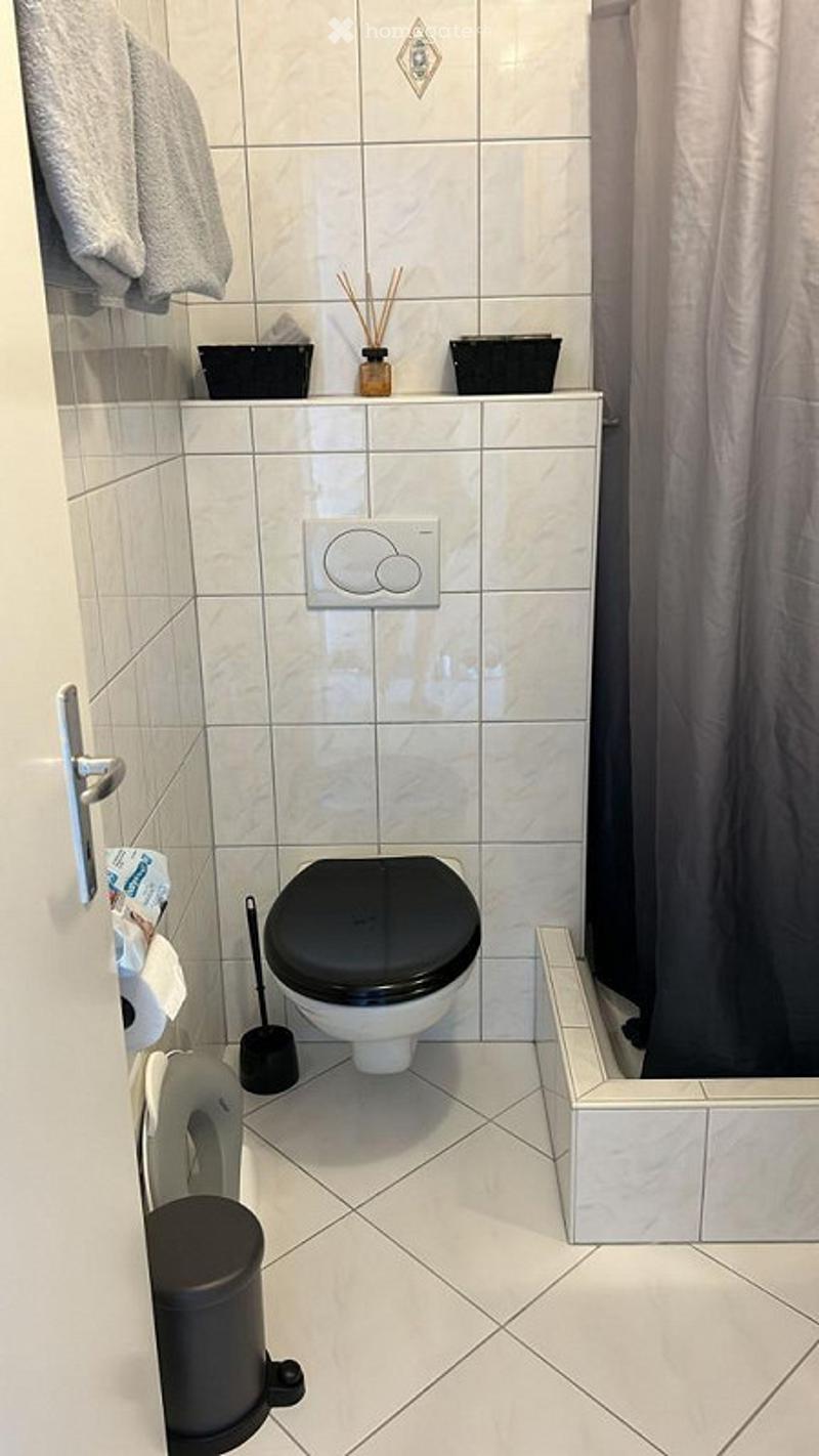 Modern bathroom with white tiled walls and floor, black toilet seat, towel hanging on the wall, toilet paper holder, and decorative items on the shelf.