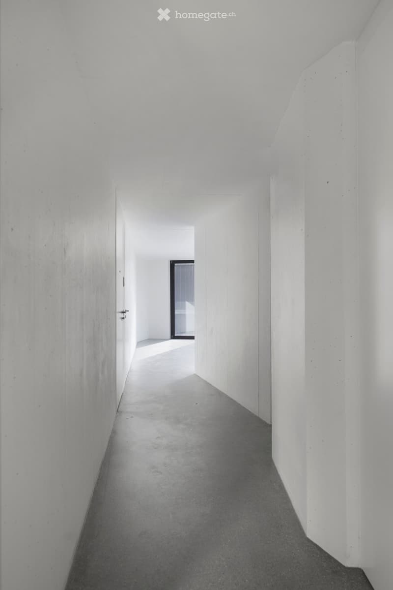 A long, narrow hallway with white walls and a concrete floor, leading to an open doorway at the end.