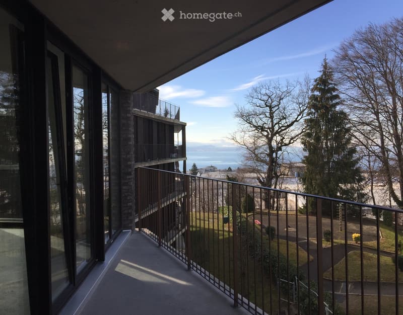 Balcony with metal railing, overlooking a landscaped area with trees and a body of water in the distance.