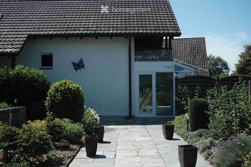 A well-maintained garden with a paved pathway leading to a modern house entrance. The garden features neatly trimmed bushes, potted plants, and a decorative butterfly sculpture on the wall.