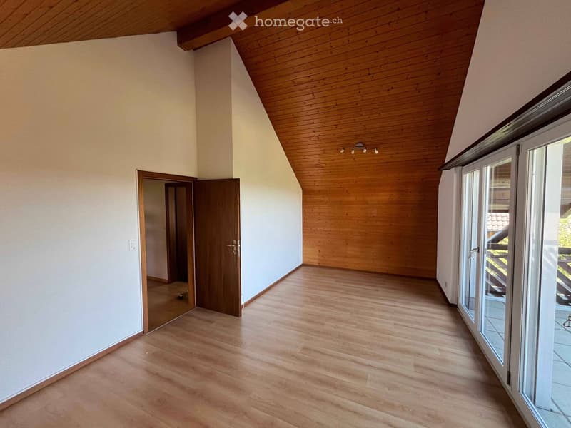 Empty room with wooden flooring, white walls, and a slanted wooden ceiling. Features a large window and a door leading to another room.